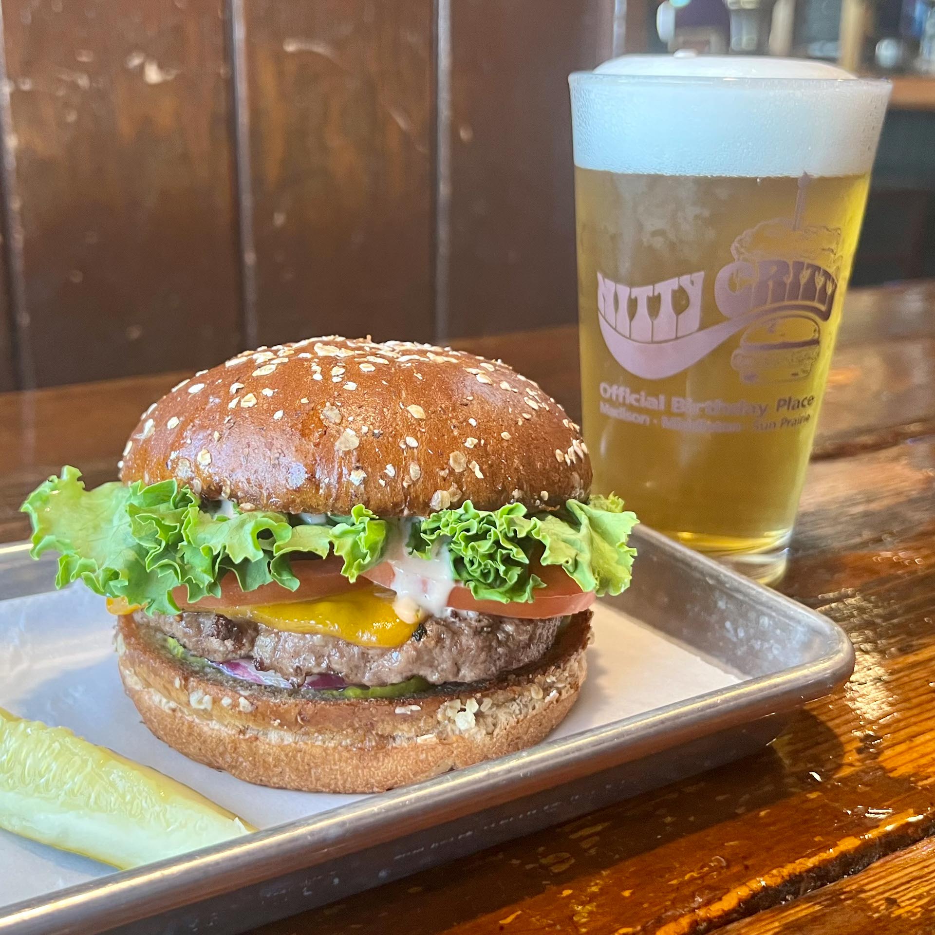 A cheeseburger with lettuce, tomato, and sauce on a wheat bun sits on a tray next to a pickle spear and a pint of beer on a wooden table.