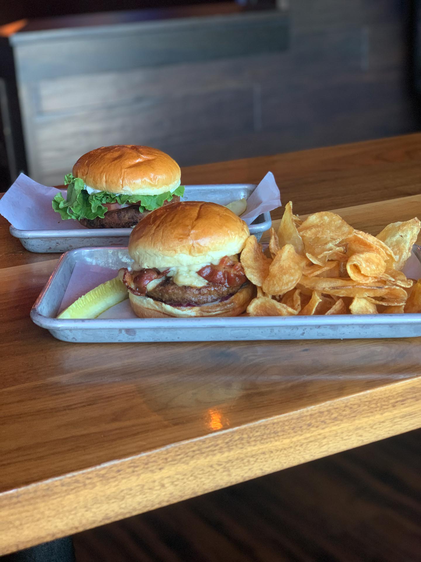 Two metal trays on a wooden table, each with a burger on a bun; one tray also has potato chips and a pickle spear.
