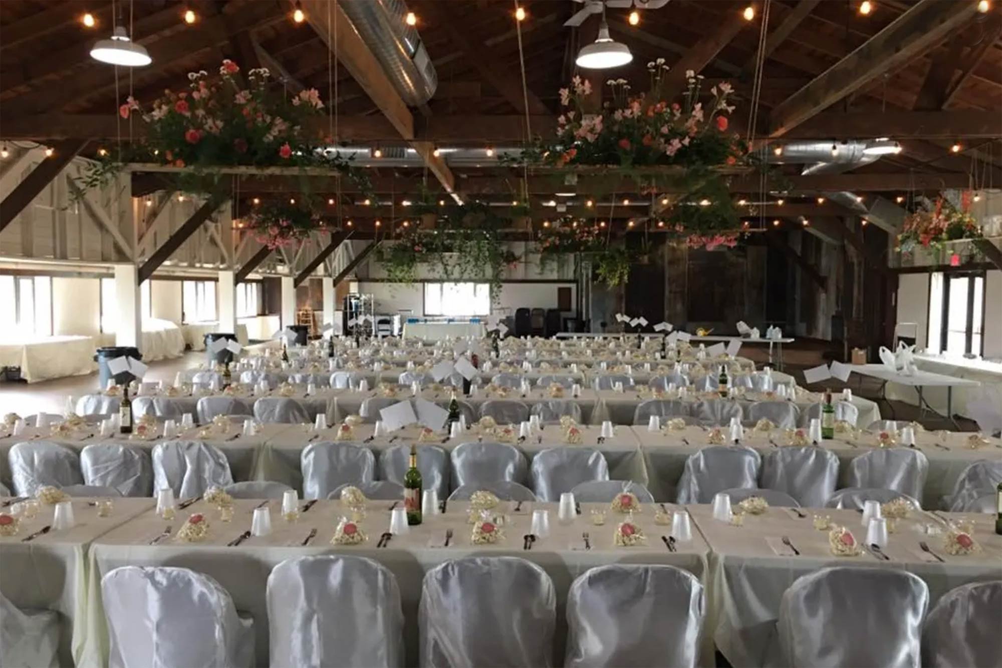 Rows of banquet tables with white tablecloths and covered chairs set for an event in a rustic hall with string lights and floral arrangements hanging from the ceiling.