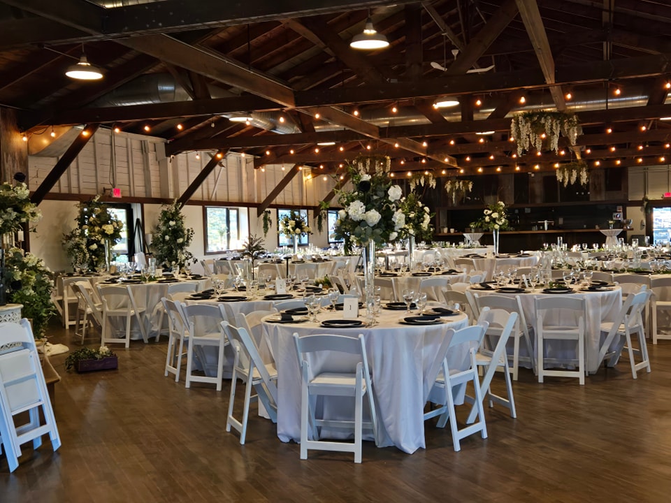 A decorated banquet hall with round tables covered in white tablecloths, white chairs, floral centerpieces, and string lights hanging from the wooden ceiling.