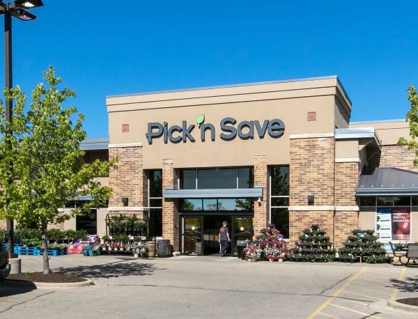 Pick 'n Save grocery store exterior with brick façade, large sign above entrance, potted plants and flowers displayed outside, and clear blue sky in the background.