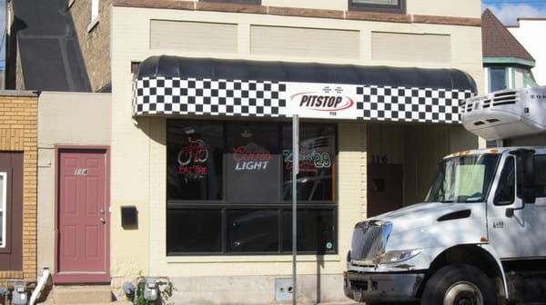 A small storefront with a checkered awning reading "Pitstop Pub" and neon beer signs in the window; a white truck is parked in front.