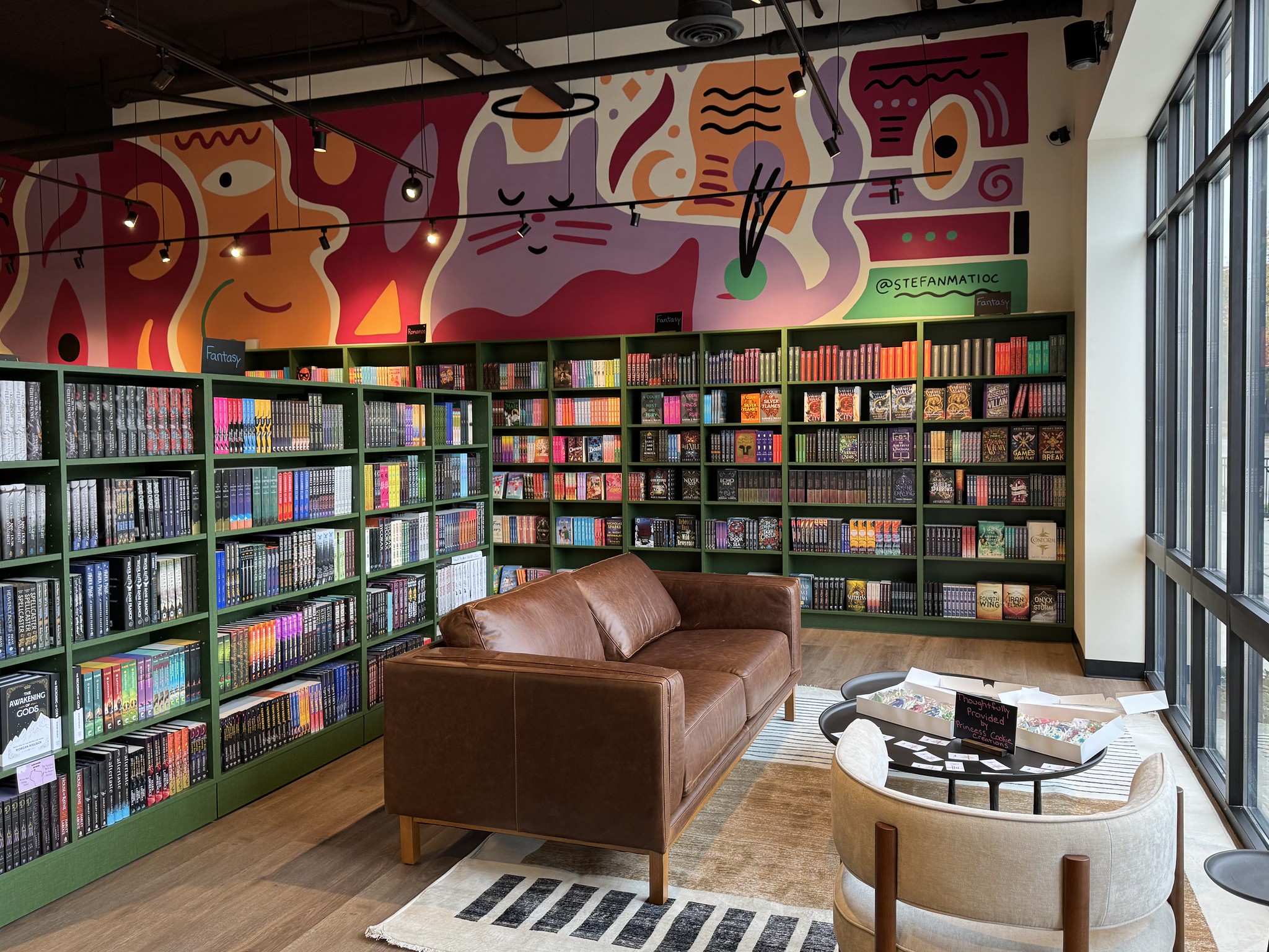 A cozy bookstore lounge with a brown leather sofa, bookshelves filled with colorful books, a modern mural on the wall, and large windows letting in natural light.