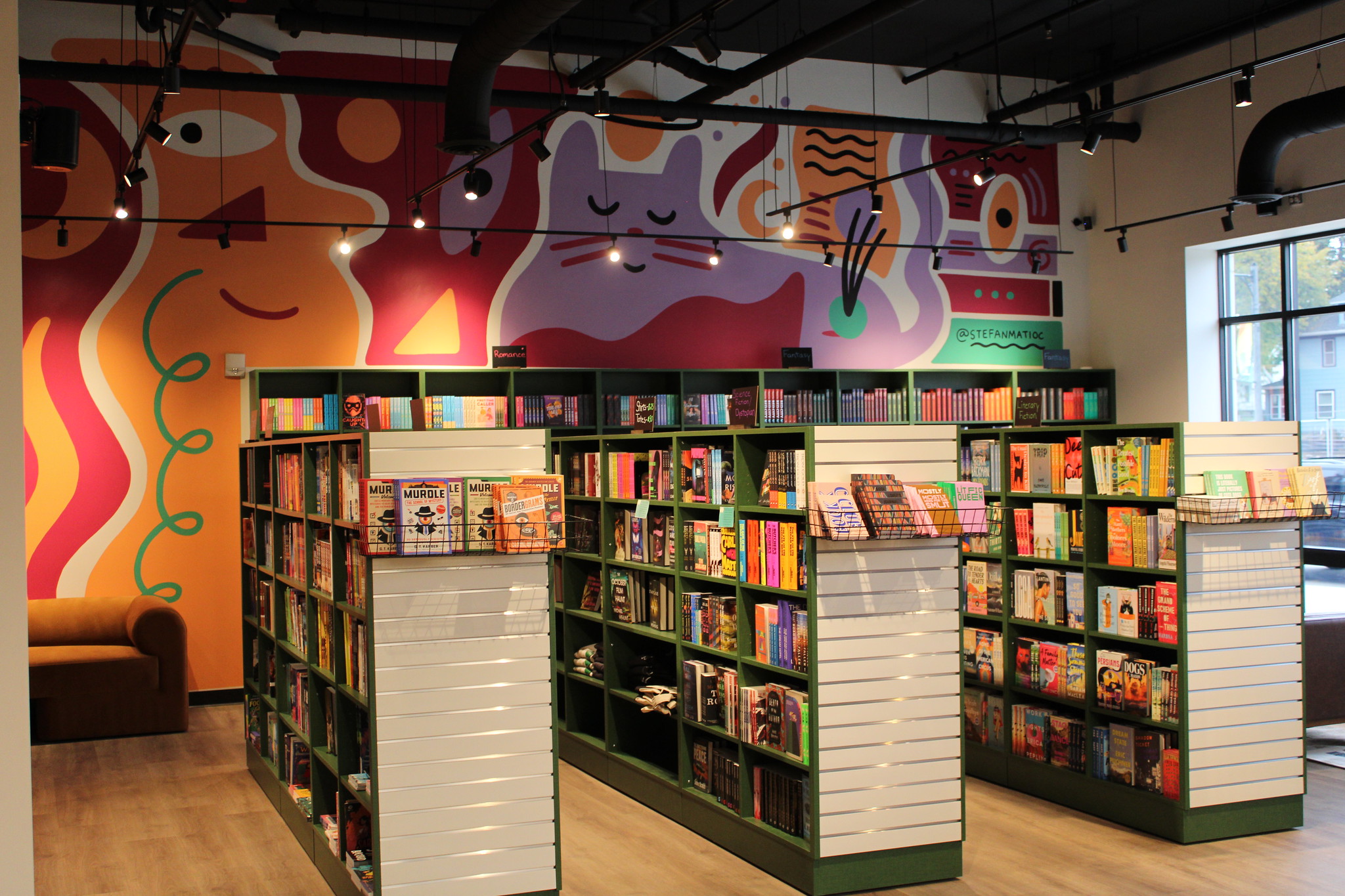 Bookshelves filled with colorful books in a bookstore, with a large, vibrant mural featuring abstract animal shapes on the back wall.