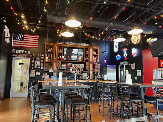 Casual bar interior with high tables, bar stools, American flag, string lights, wall decorations, and a stocked bar counter in the background.