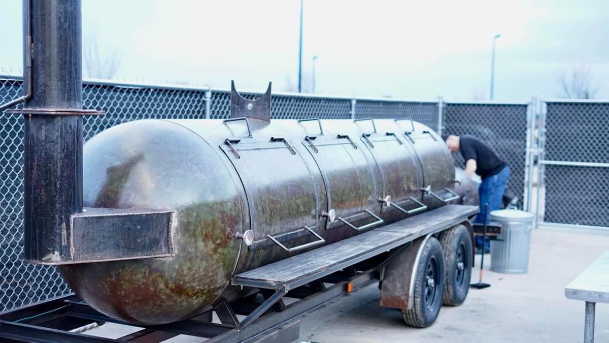 Large metal barbecue smoker mounted on a trailer, situated outdoors near a chain-link fence, with a person working beside it.