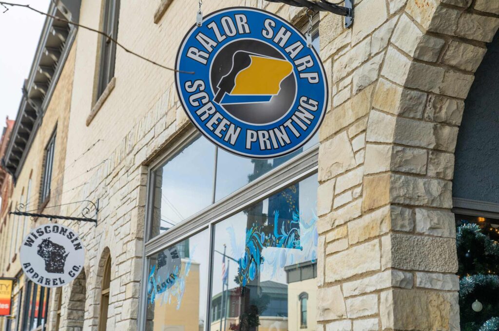 A round blue sign for "Razor Sharp Screen Printing" hangs above a shop entrance on a stone building in a city setting.