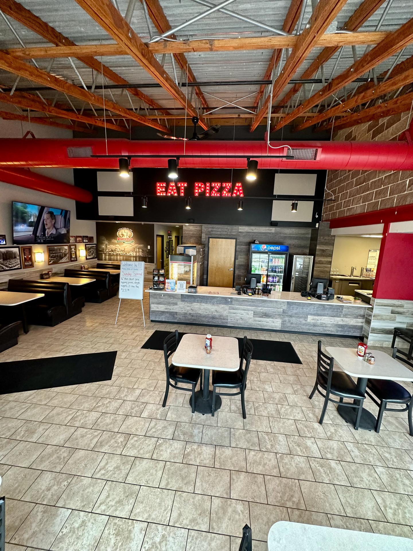 Restaurant interior with tiled floor, exposed ceiling, tables and booths, a counter with soda machines, and a lit "EAT PIZZA" sign above the counter.