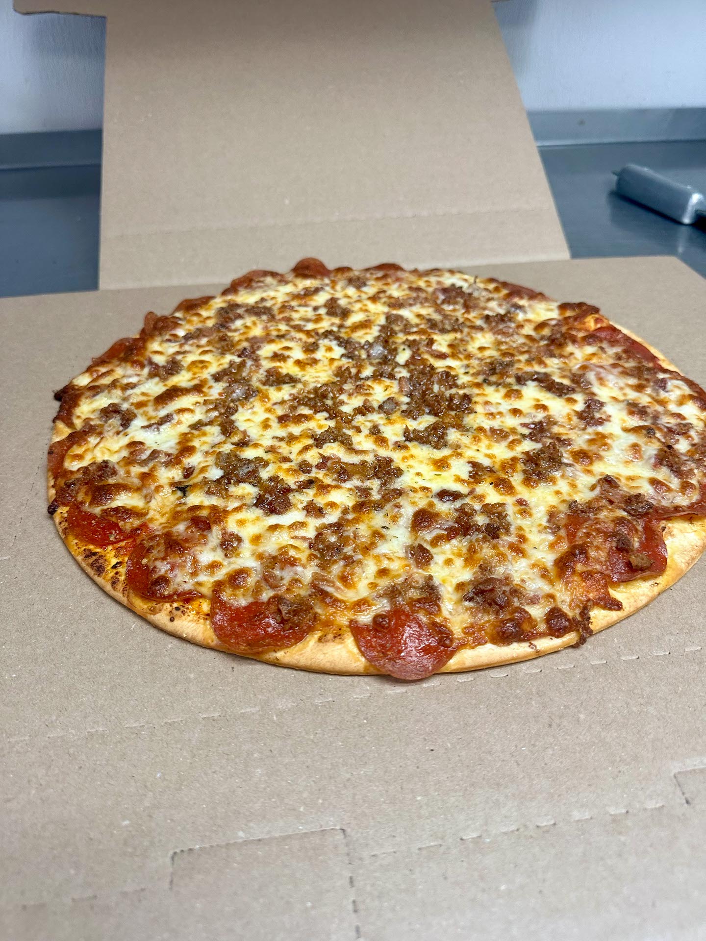 A cooked pizza with a golden, slightly crispy crust, topped with melted cheese and pieces of meat, sits on an open cardboard pizza box.