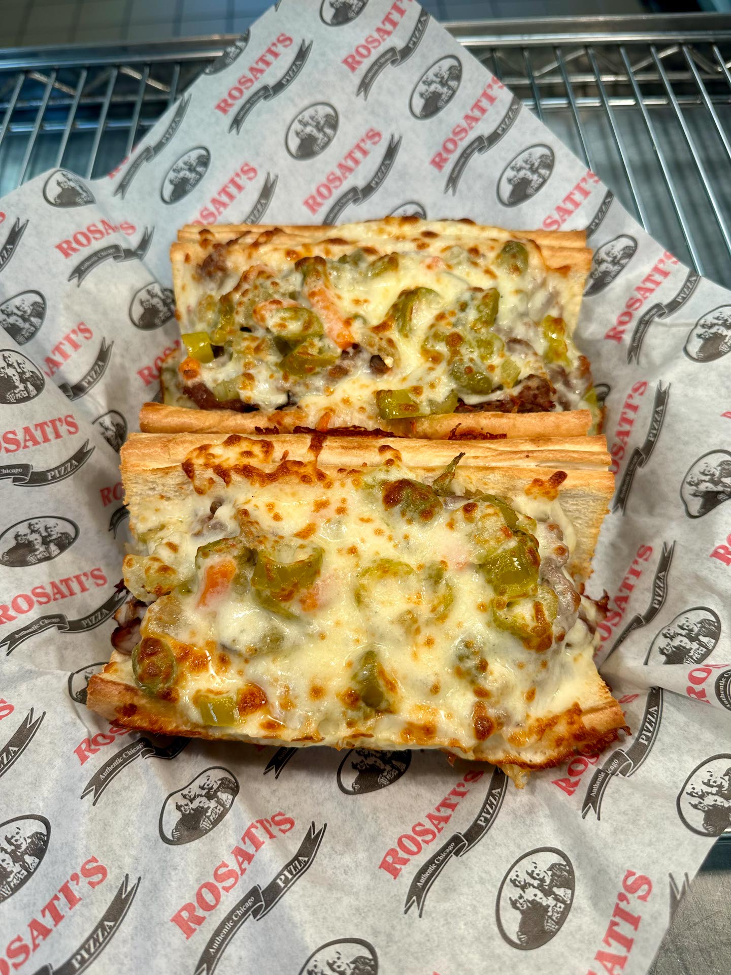 Two pieces of cheesy bread topped with chopped green peppers, onions, and melted cheese on Rosati’s branded paper in a metal basket.
