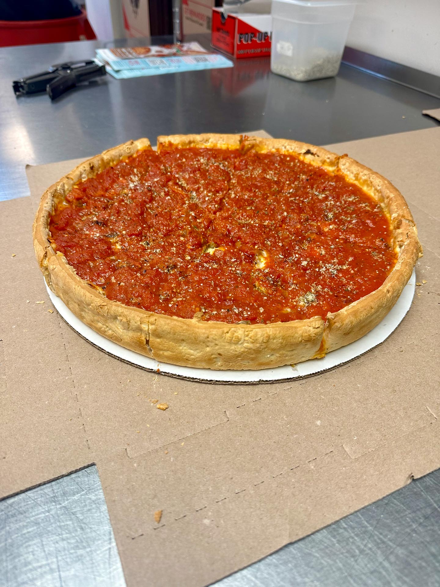 A whole Chicago-style deep dish pizza with a thick crust and tomato sauce sits on a cardboard base on a stainless steel counter.