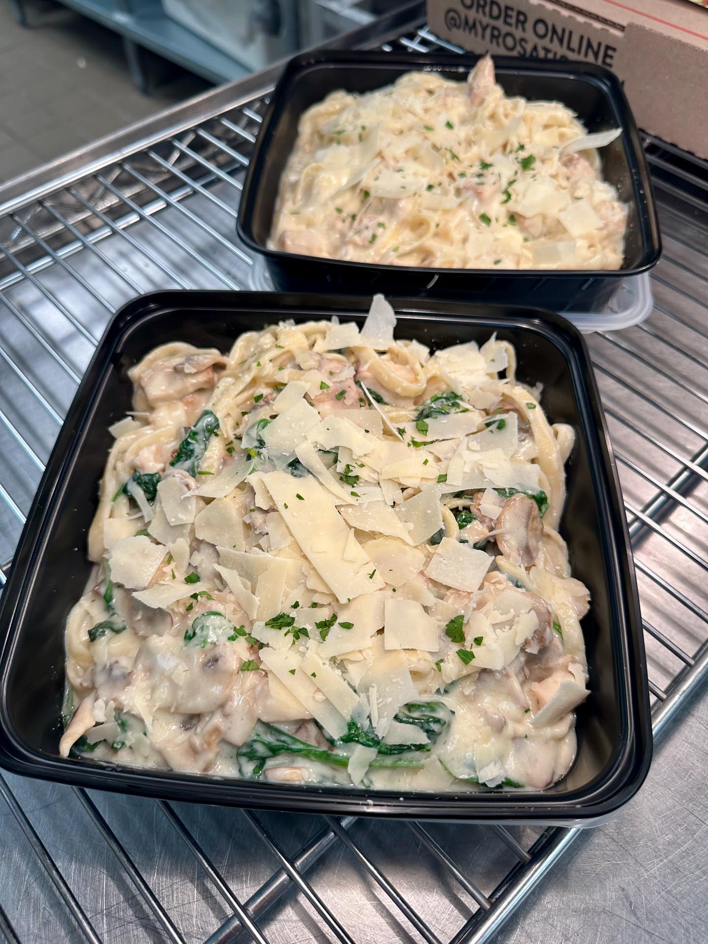 Two black plastic containers filled with fettuccine in creamy Alfredo sauce, garnished with spinach, sliced chicken, and shaved parmesan, sit on a metal rack.