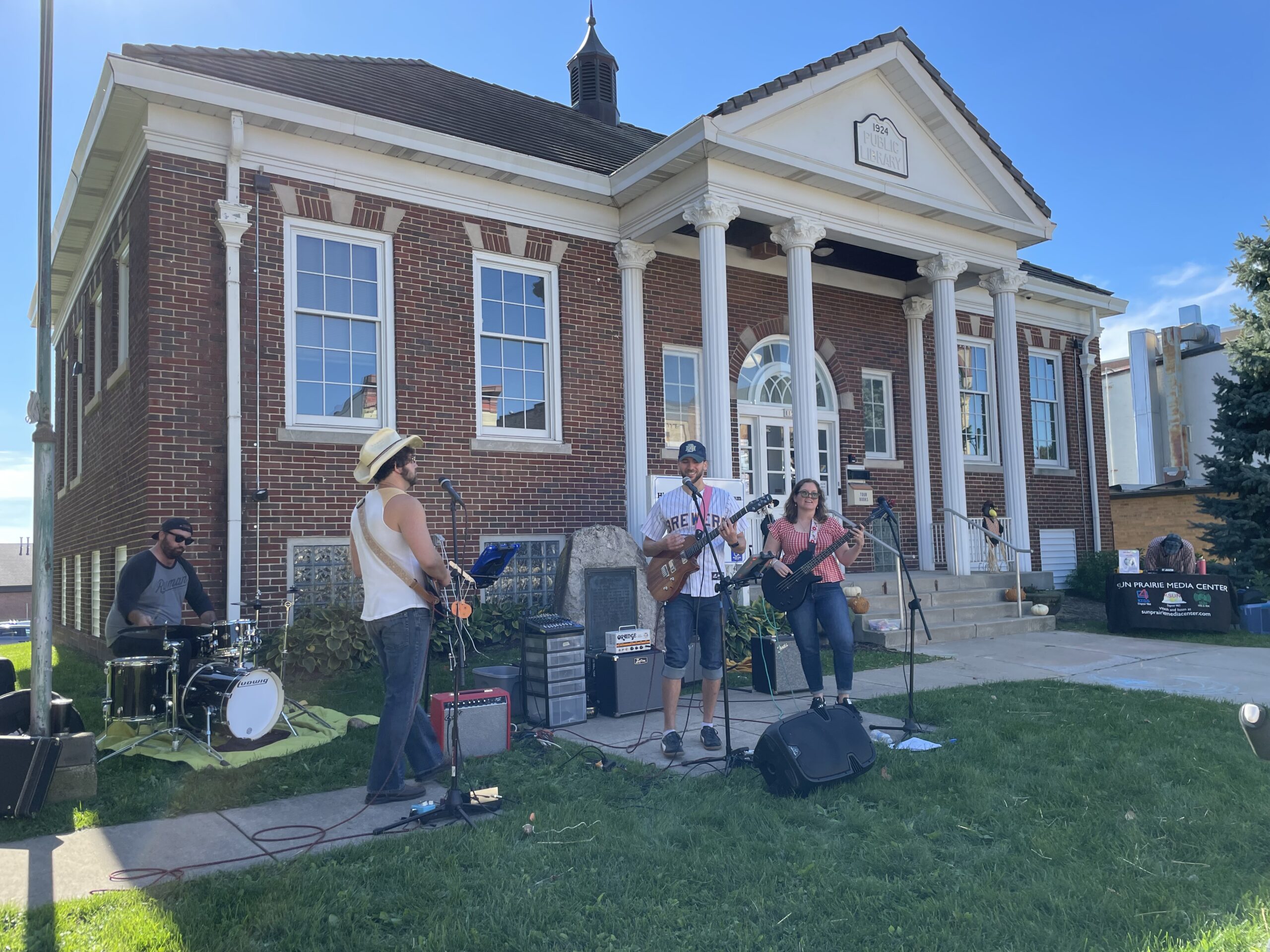 A band performs outdoors on the grass in front of a brick building with columns while people watch and a person sits at a table nearby.