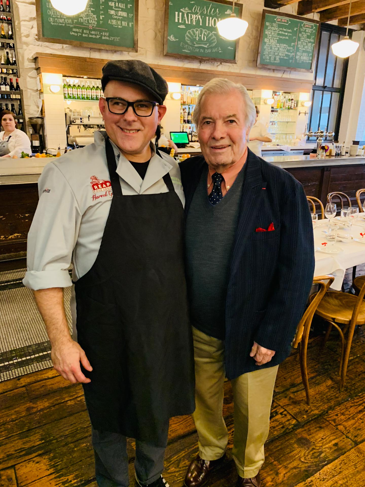 Two men standing and smiling together inside a restaurant, one wearing a chef's uniform and apron, the other dressed in a suit.