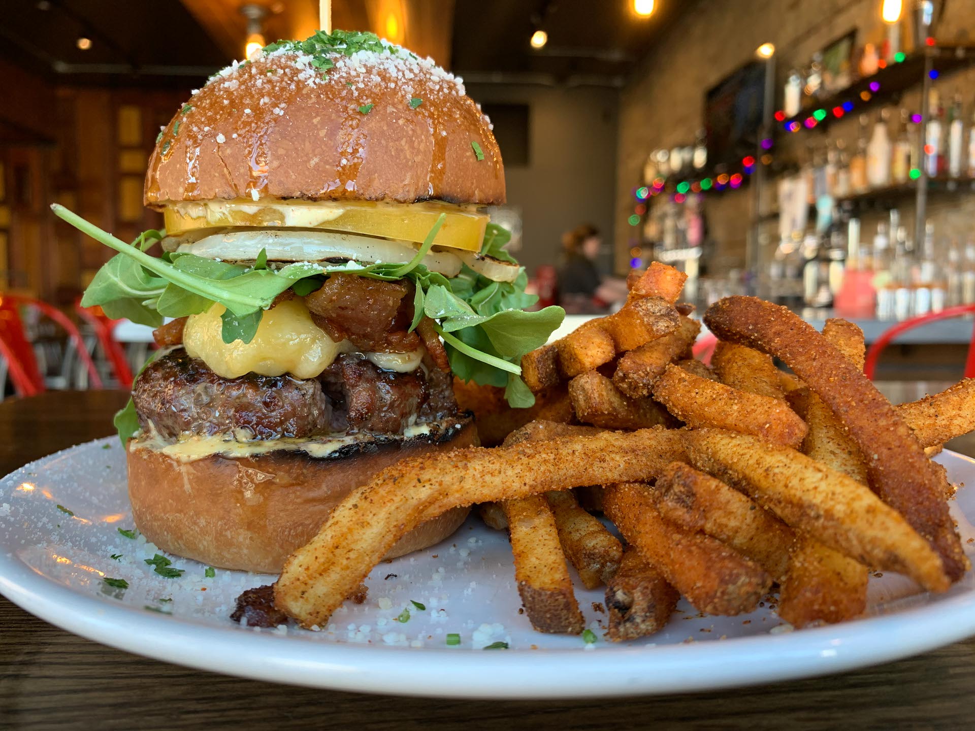 A cheeseburger with lettuce, tomato, onions, and sauce on a bun is served with seasoned French fries on a white plate in a restaurant setting.