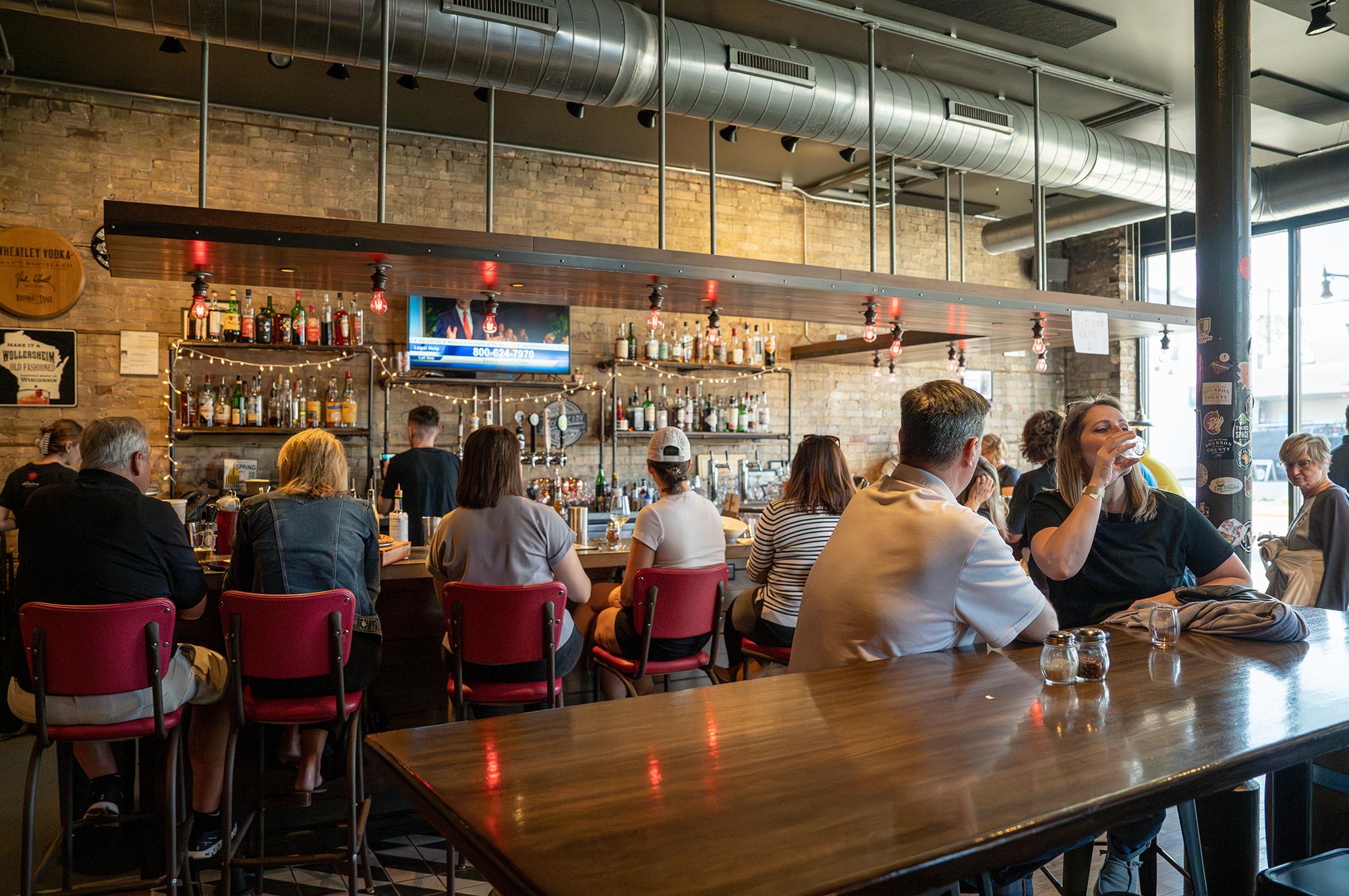 People sit at a bar and tables inside a modern, industrial-style restaurant with exposed brick walls and a TV above the bar.