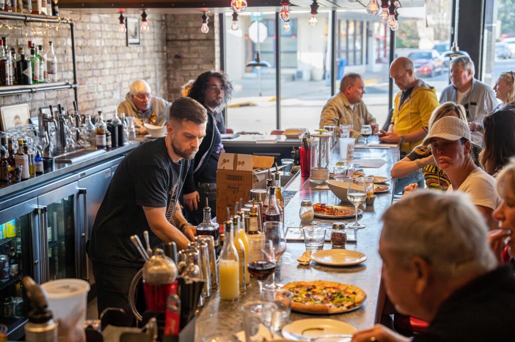 Bartender working behind a busy bar as patrons eat and drink at the counter; pizzas and drinks are visible on the countertop.