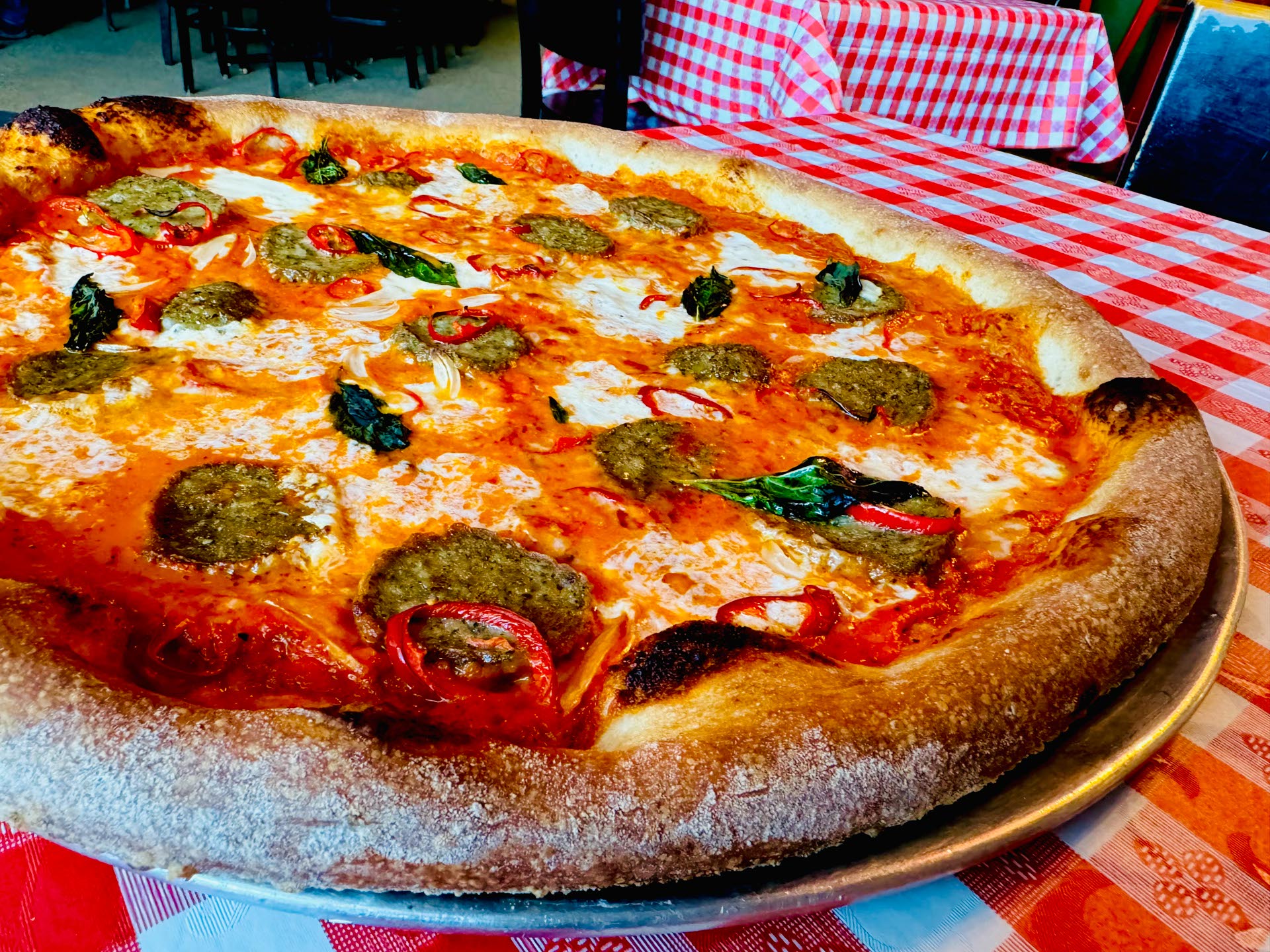 A large pizza with melted cheese, tomato sauce, basil leaves, and sausage slices sits on a metal tray atop a red and white checkered tablecloth.