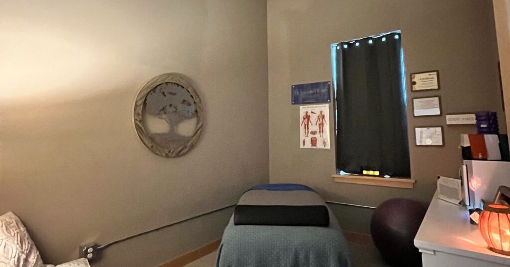 A small therapy room with a massage table, a window with a dark curtain, anatomical charts on the wall, and soft lighting.