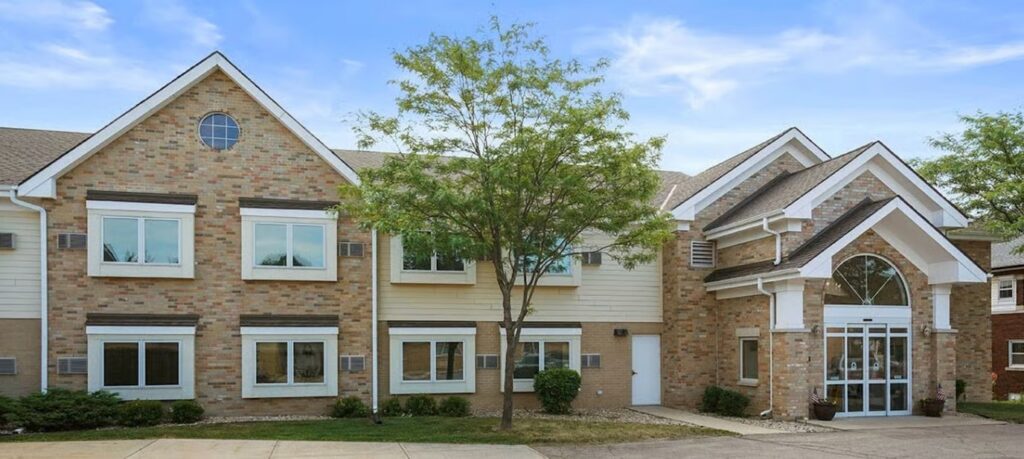 Two-story brick building with white trim and large front entrance, featuring multiple windows and a small tree in front.