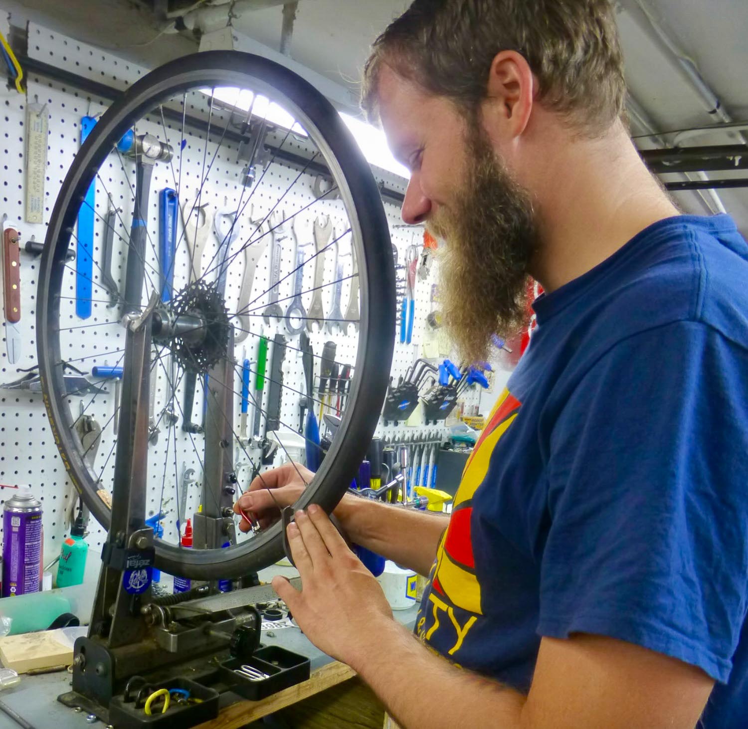 A man uses a truing stand to adjust the spokes of a bicycle wheel in a workshop with tools hanging on the wall.