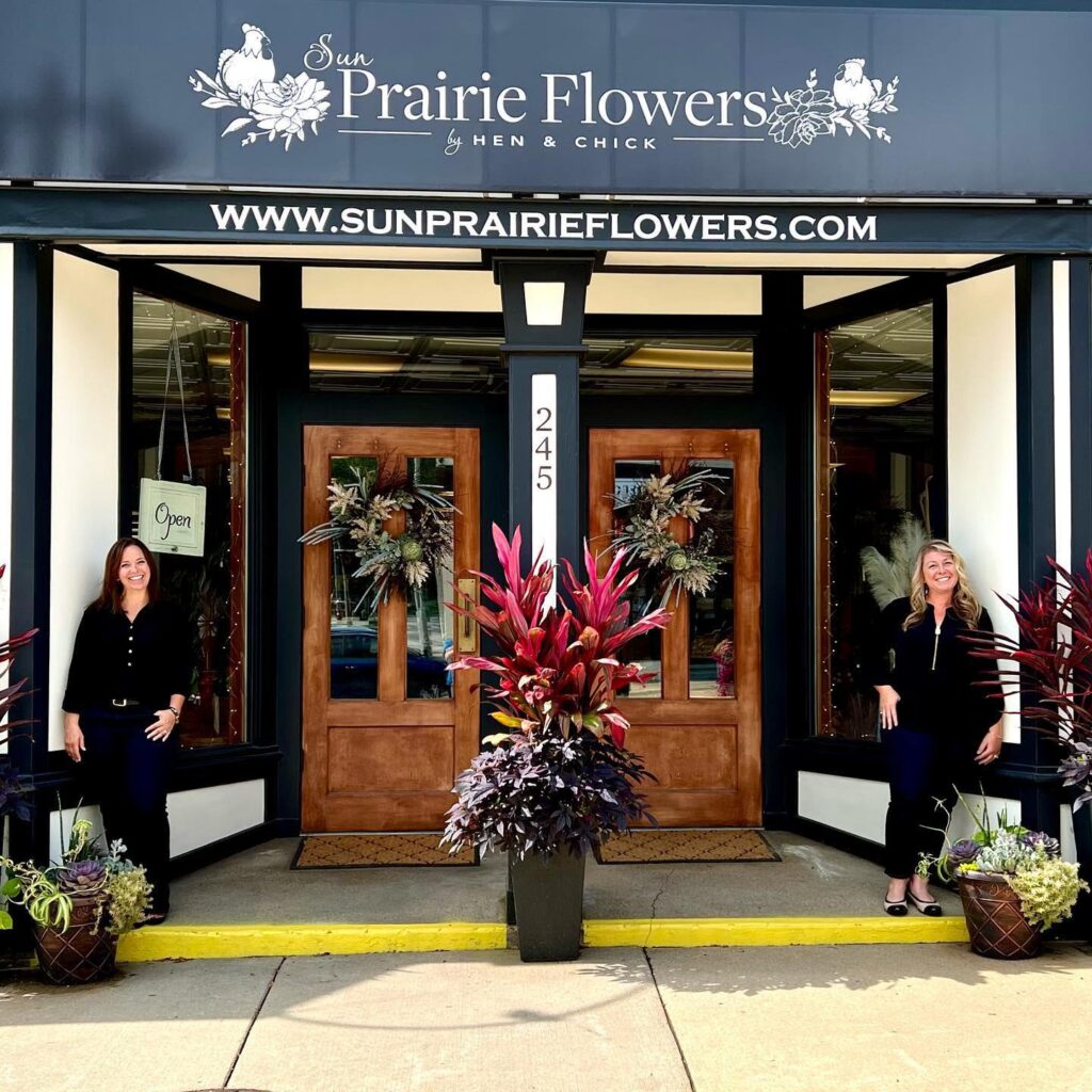 Two women stand on either side of the entrance to Sun Prairie Flowers, which has double wooden doors decorated with floral wreaths and potted plants in front.