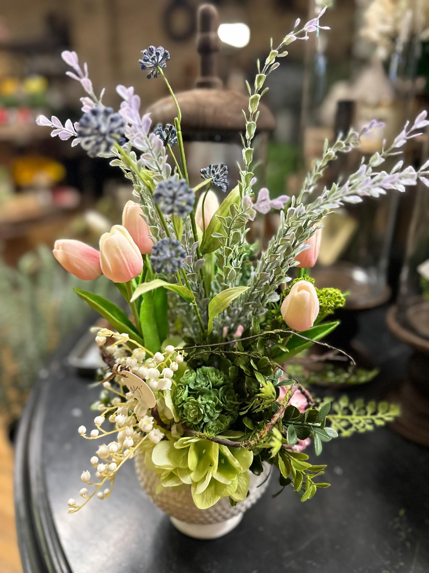 A small white vase filled with pink tulips, lavender, green succulents, and assorted greenery sits on a dark tabletop.