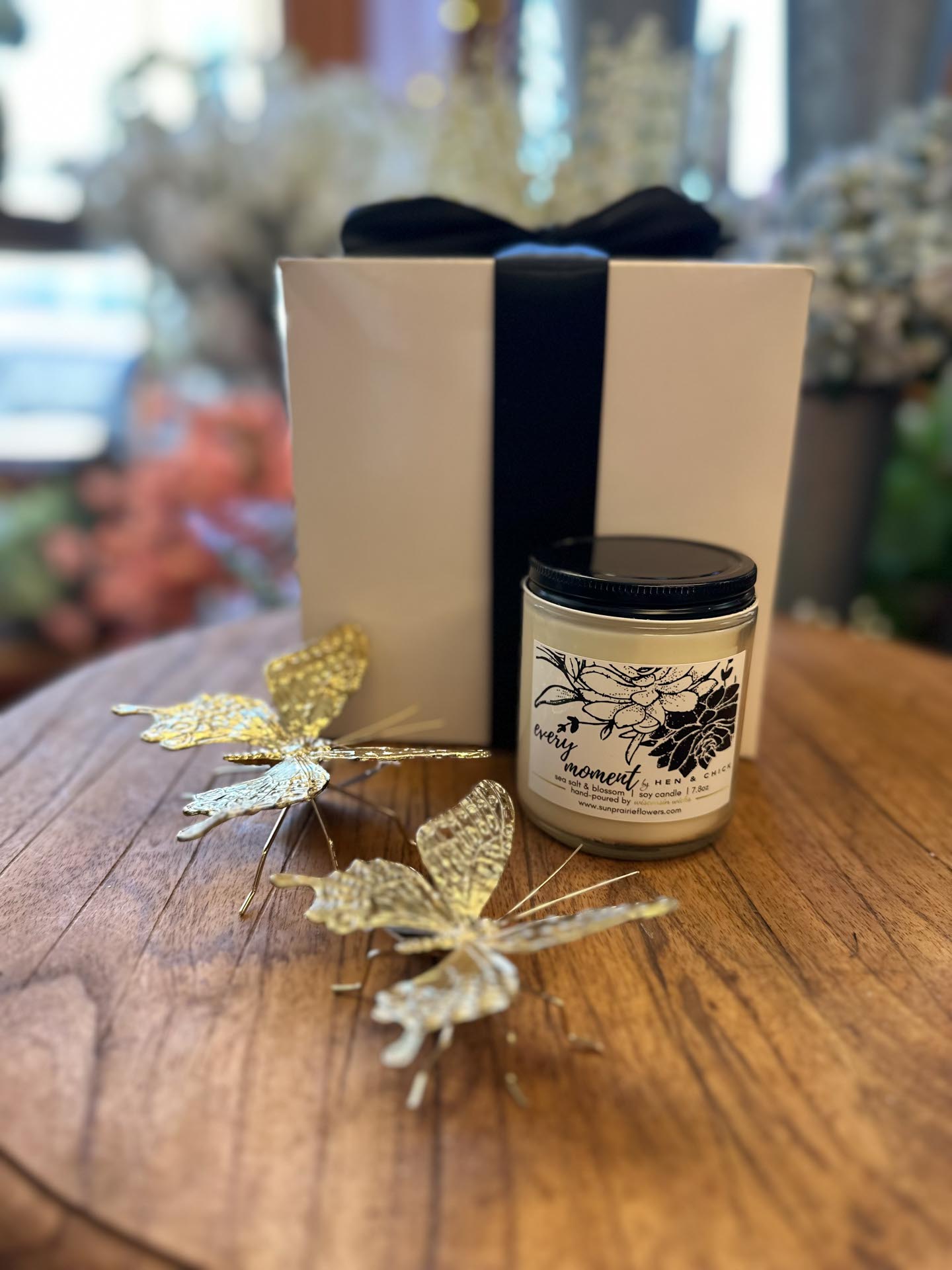 A white gift box with a dark blue ribbon and a candle jar sit on a wooden table, beside two decorative gold butterflies.