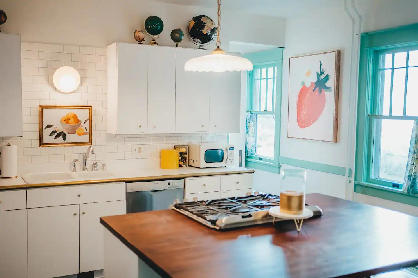 Bright kitchen with white cabinets, a wooden island with a stove, green-framed windows, and fruit-themed artwork on the walls. A globe sits on top of the cabinets.