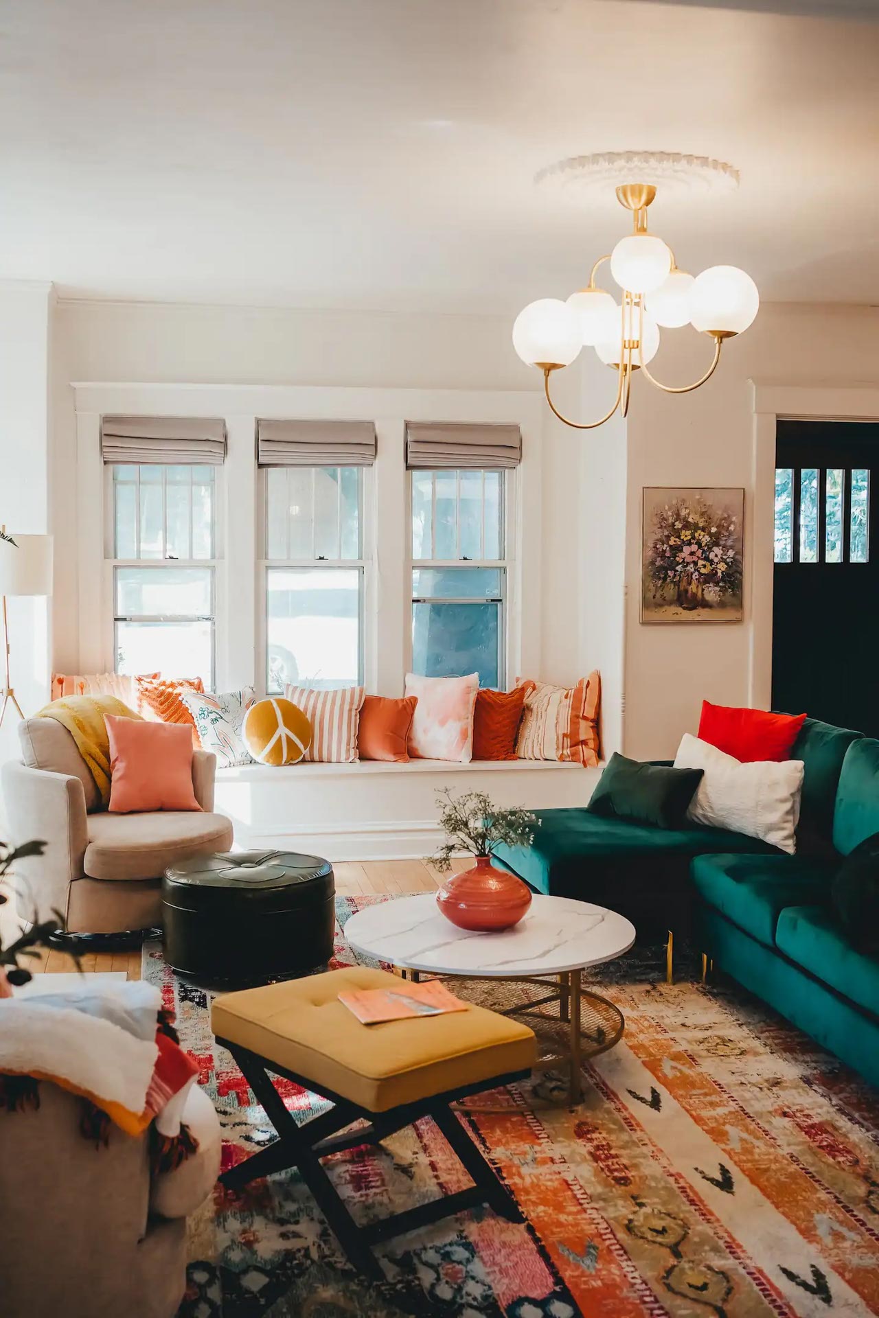 A brightly lit living room with a green sofa, colorful cushions, a marble coffee table, a large rug, a bench, and a window seat under three windows.