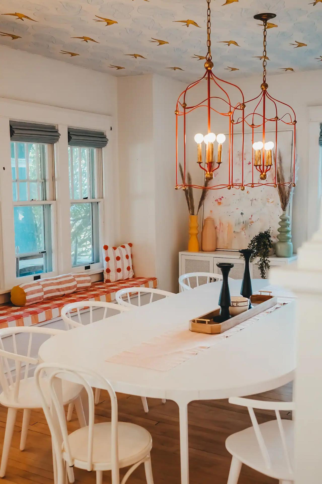 A dining room with a white table, white chairs, red and gold chandeliers, patterned bench seating, and decorative vases against a white wall.