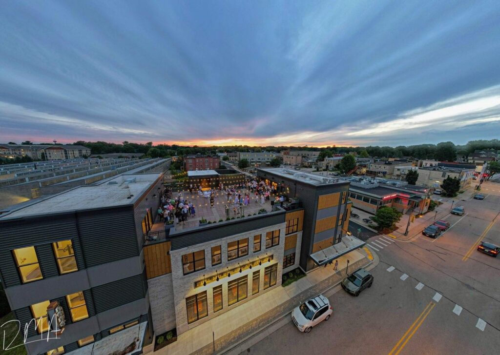A rooftop event with many people is taking place on a modern building at sunset, surrounded by city streets and other buildings.