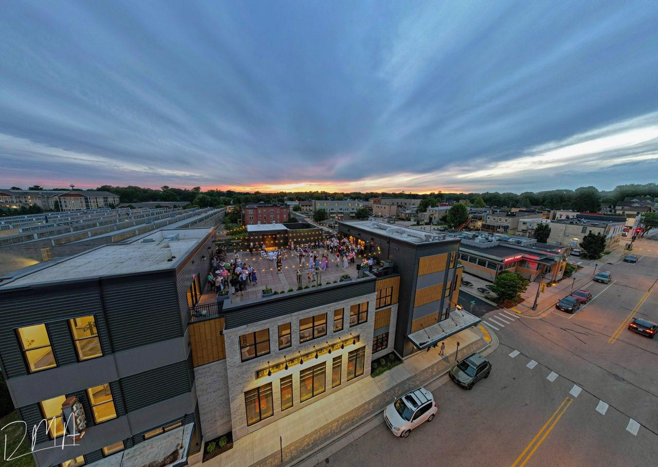 A rooftop event with many people is taking place on a modern building at sunset, surrounded by city streets and other buildings.
