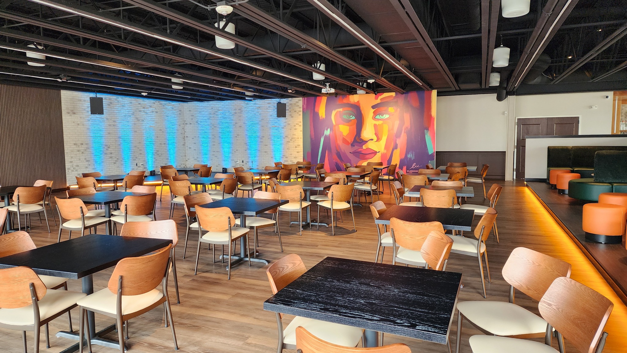 Spacious dining area with wooden tables and chairs, modern lighting, and a colorful mural of a woman's face on the back wall.