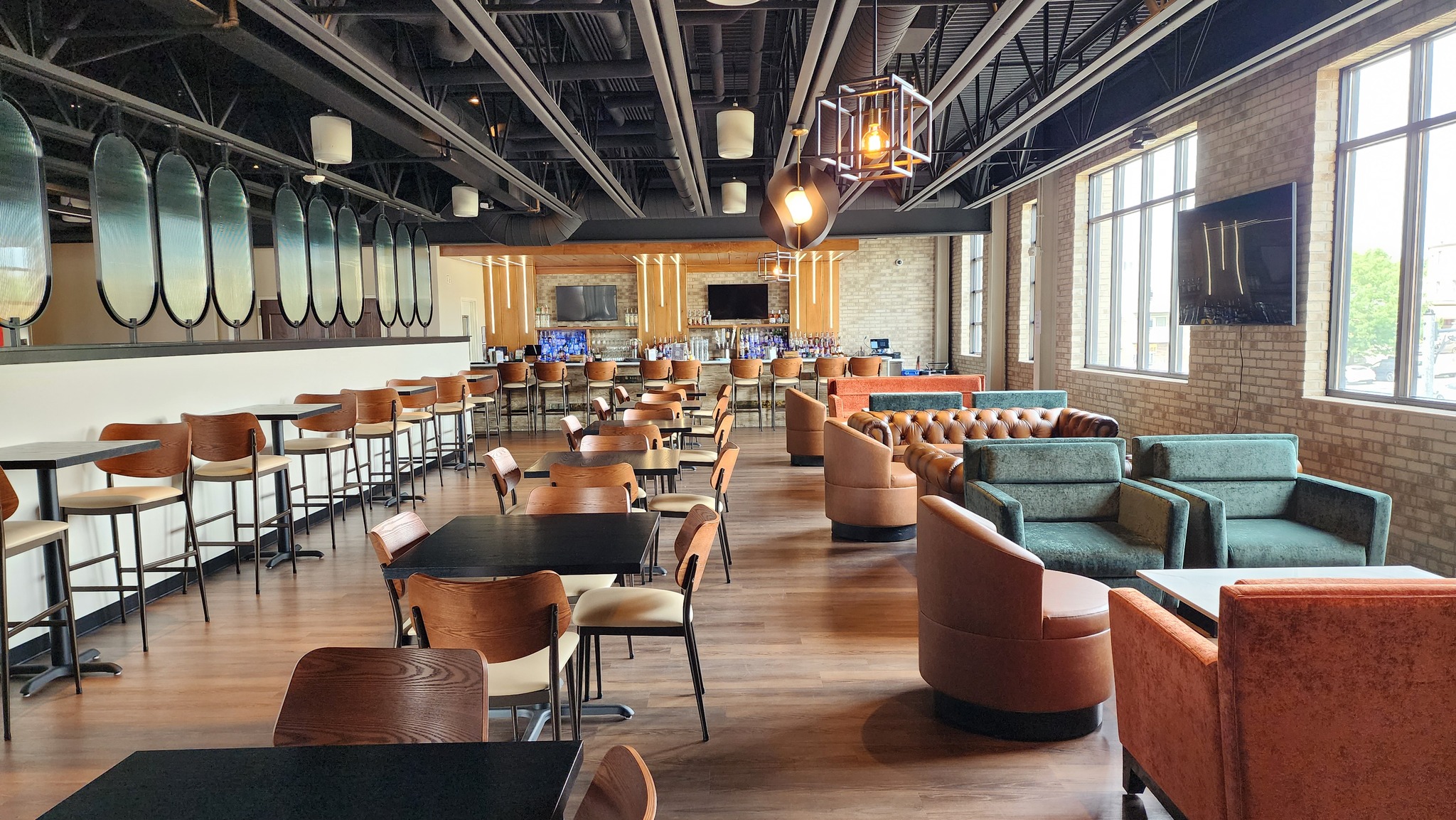 Modern restaurant interior with wooden floors, tables, and chairs, upholstered sofas, a bar area with stools, large windows, and decorative lighting.