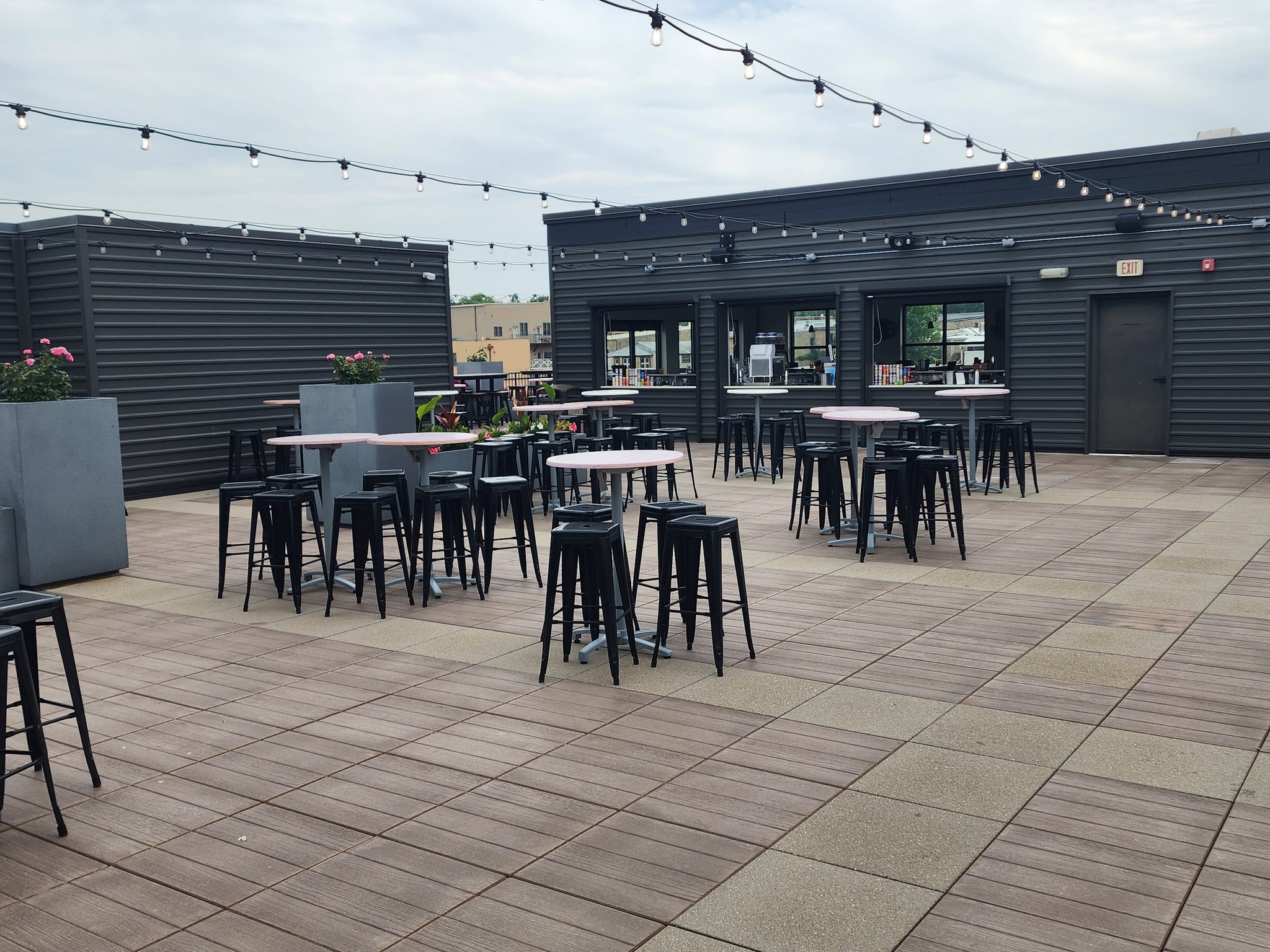 Outdoor rooftop patio with high tables and black stools, string lights overhead, and modern gray buildings along the perimeter.