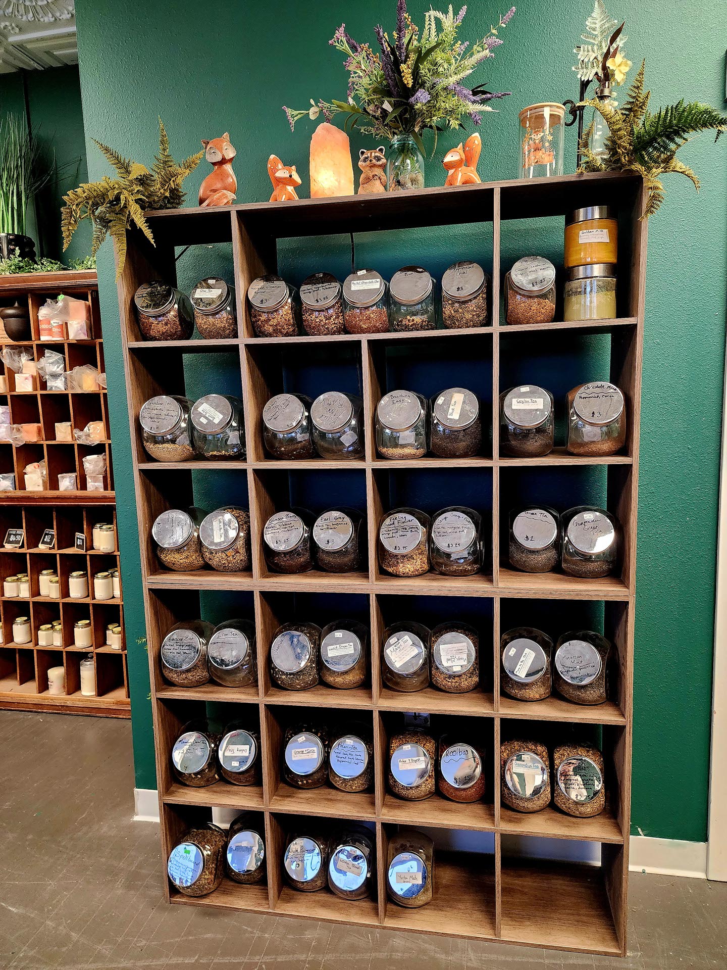 A wooden shelf with glass jars filled with herbs or tea, each labeled, stands against a green wall with decorative plants and figurines on top.