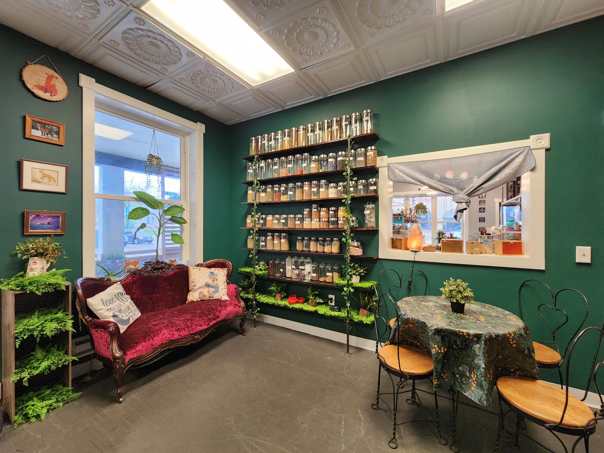 A cozy room with green walls features shelves of jars, a vintage red sofa, a small round table with chairs, and framed wall art. A window and a large interior wall opening are also visible.