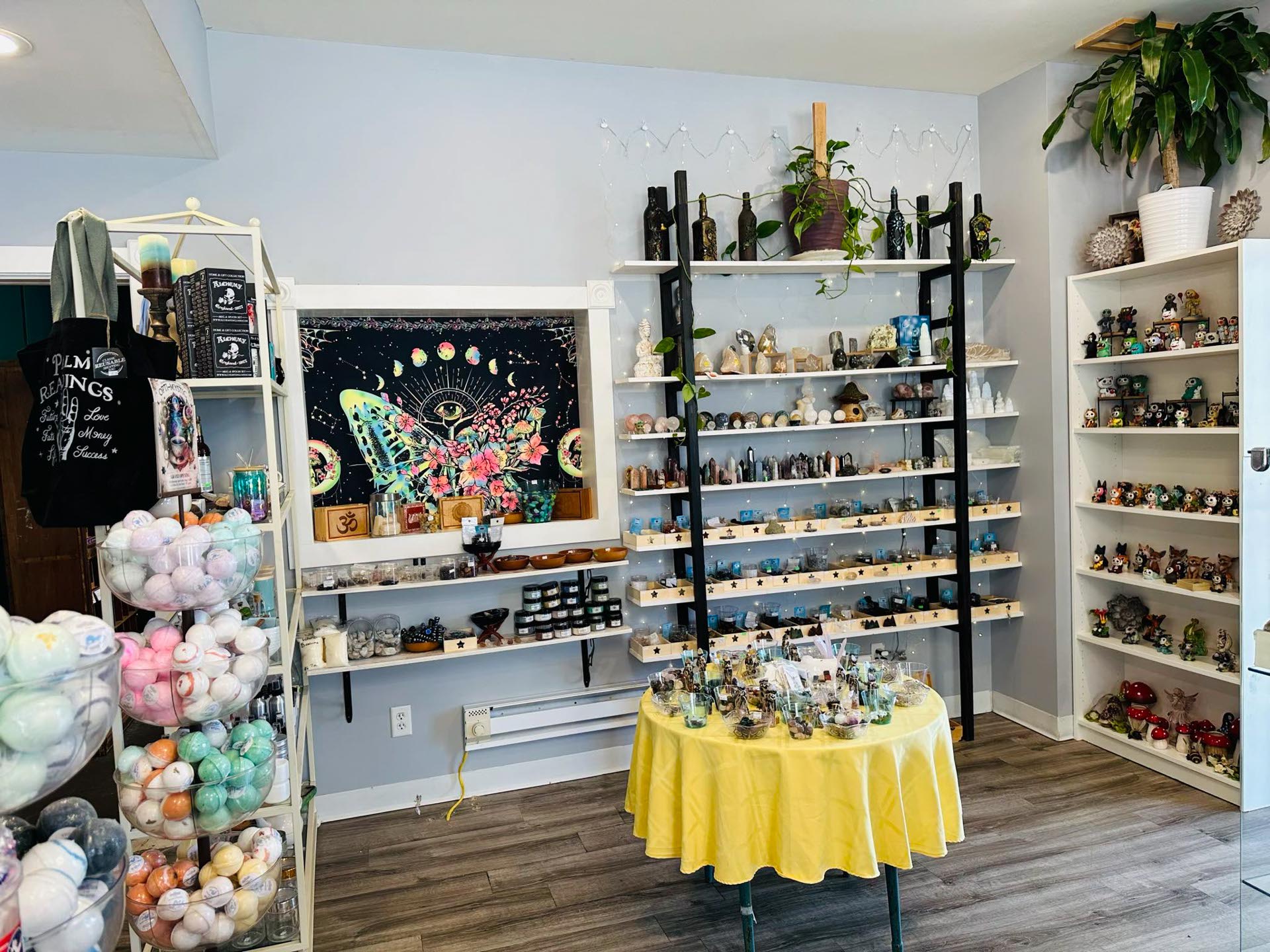 A small shop interior displays shelves filled with crystals, figurines, bath bombs, and decorative items; a round table with more items stands in the center.