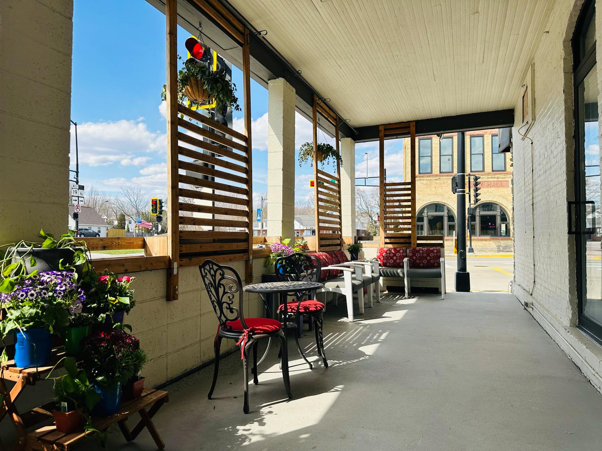 Outdoor patio with potted plants, red cushioned seating, a small table with two chairs, and wooden slat dividers, overlooking a street and brick building on a sunny day.