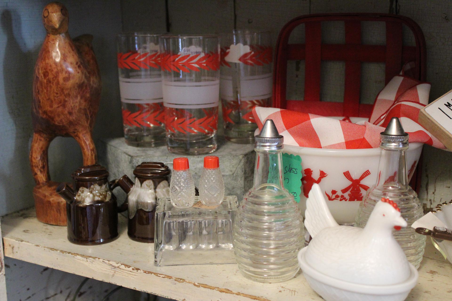 A shelf displaying vintage kitchen items, including glassware with red patterns, salt and pepper shakers, a wooden bird figurine, and a white dish with a bird lid.