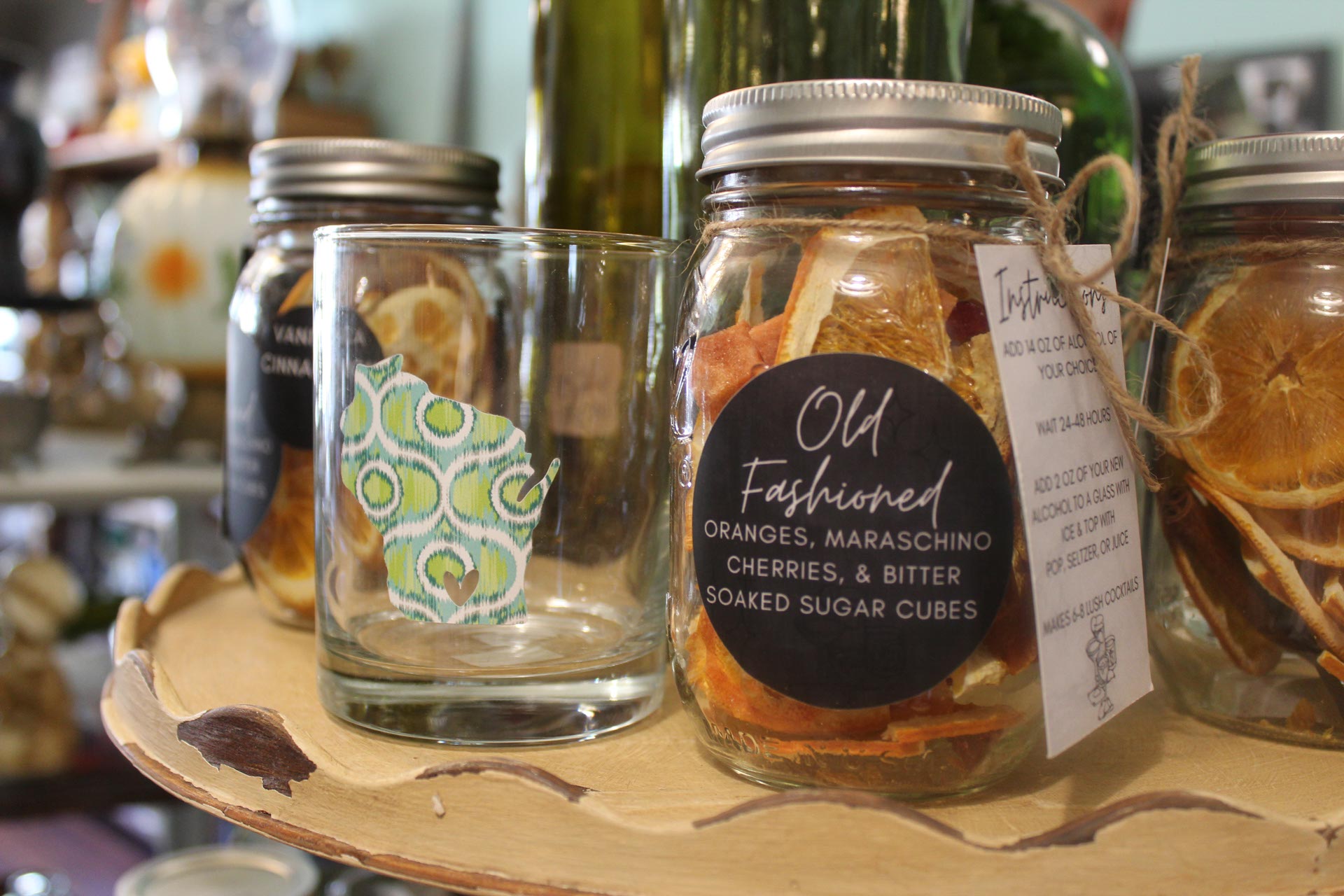 A glass with a patterned sticker next to a jar labeled "Old Fashioned" containing dried oranges, maraschino cherries, and bitter-soaked sugar cubes on a wooden tray.