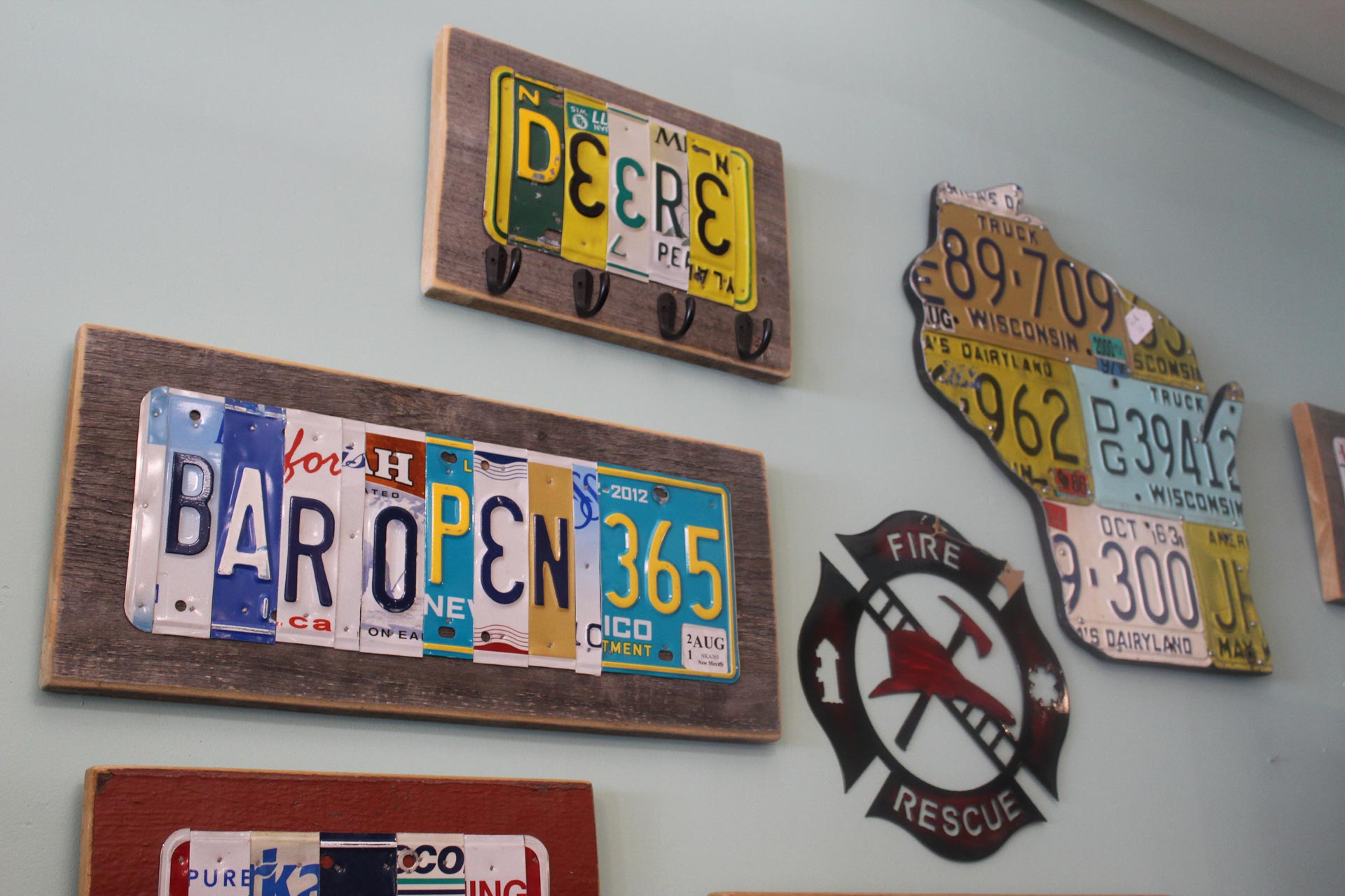 Wall art made from old license plates, including a "BAR OPEN 365" sign, a "DEERE" sign, a Wisconsin-shaped display, and a fire rescue emblem.