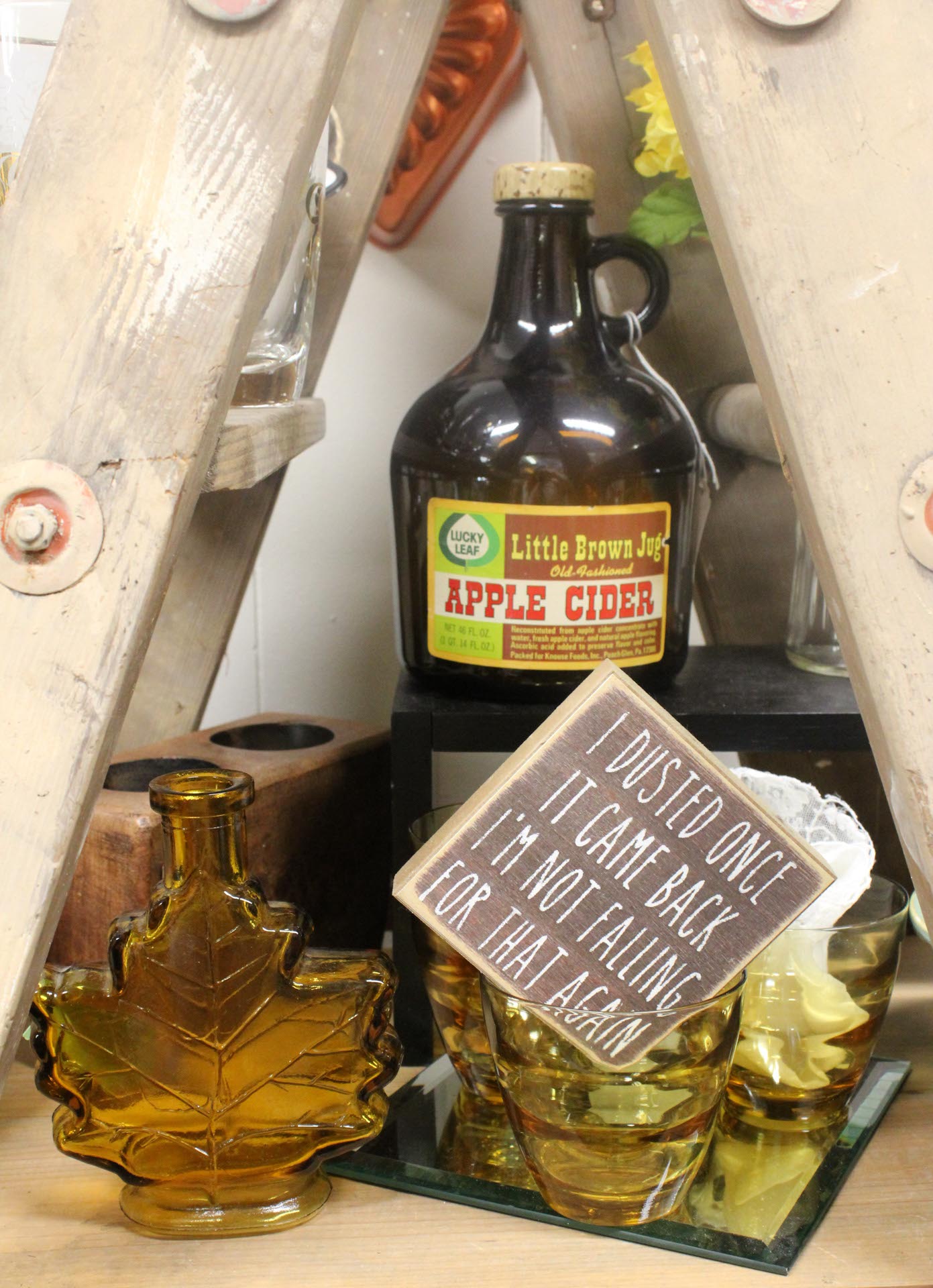 Amber glass containers, a jug labeled "Apple Cider," and a wooden sign that reads, "I dusted once, it came back, I'm not falling for that again," are displayed on a shelf.