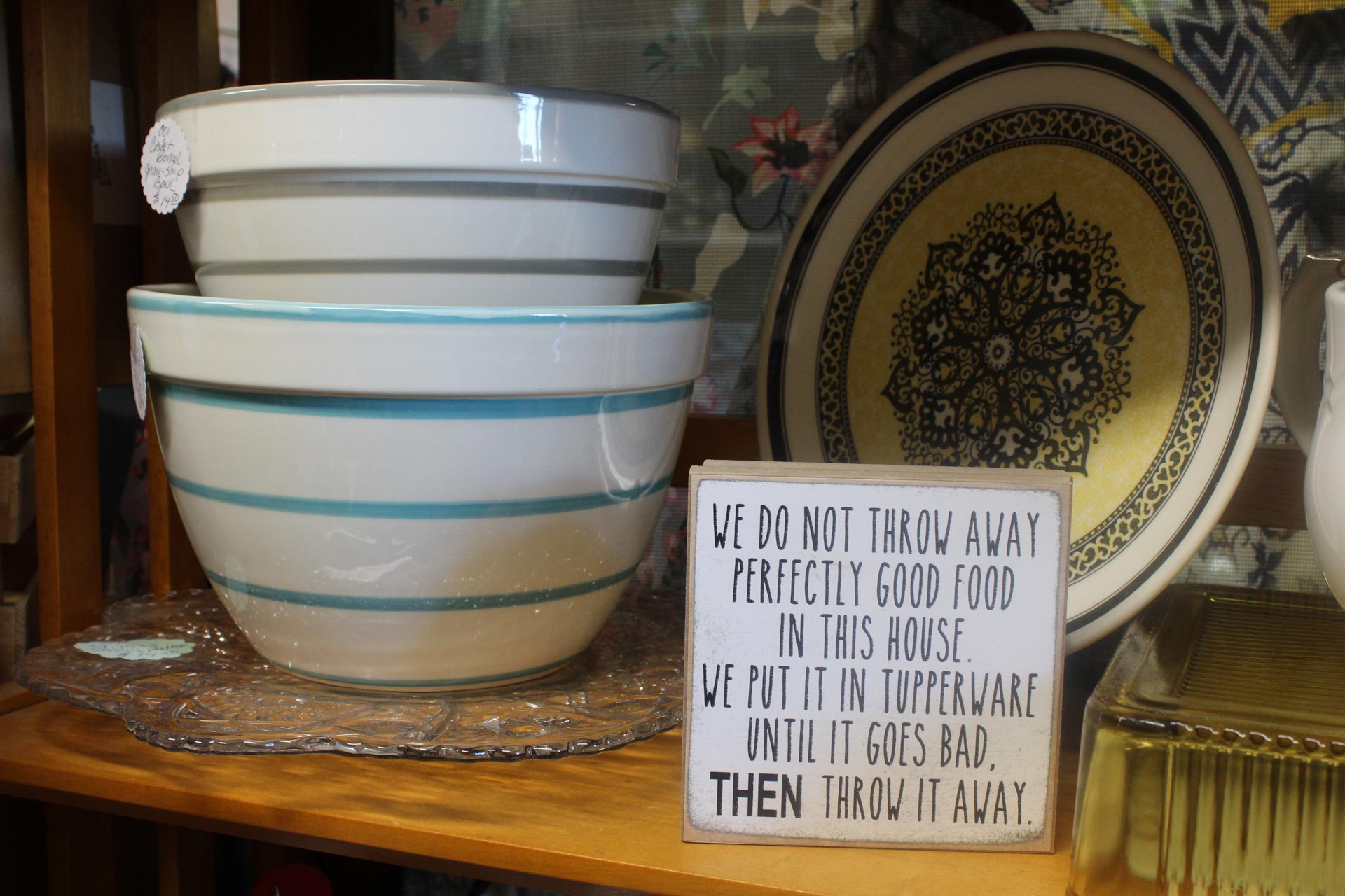 Two ceramic mixing bowls stacked next to a decorative plate and a sign that reads, "We do not throw away perfectly good food in this house..." on a wooden shelf.