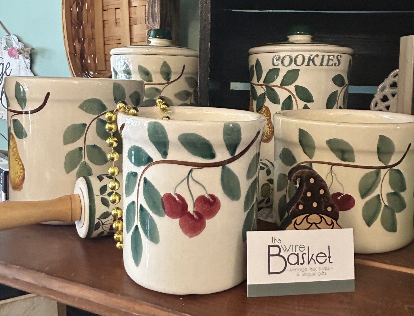 Ceramic kitchen canisters with leaf and cherry designs are displayed on wooden shelves alongside a rolling pin, beads, and a “the wire Basket” business card.