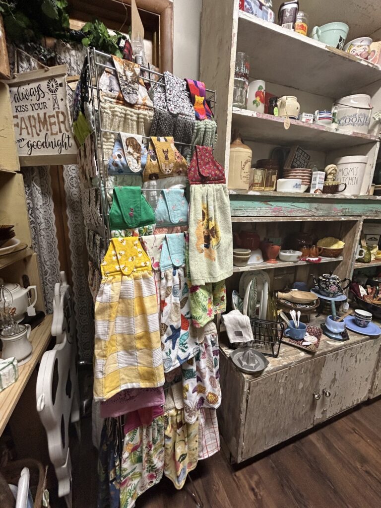 A display rack holds colorful hanging kitchen towels next to shelves filled with assorted vintage kitchenware and decor in a rustic shop setting.