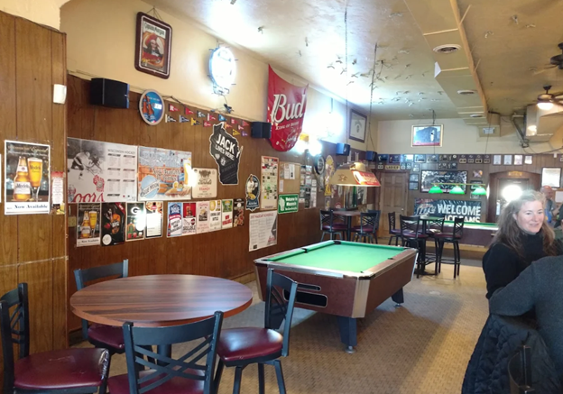 A bar interior with a pool table, round tables and chairs, posters on the wall, and a woman sitting near the front. Neon beer signs and a "Welcome" sign are visible in the background.