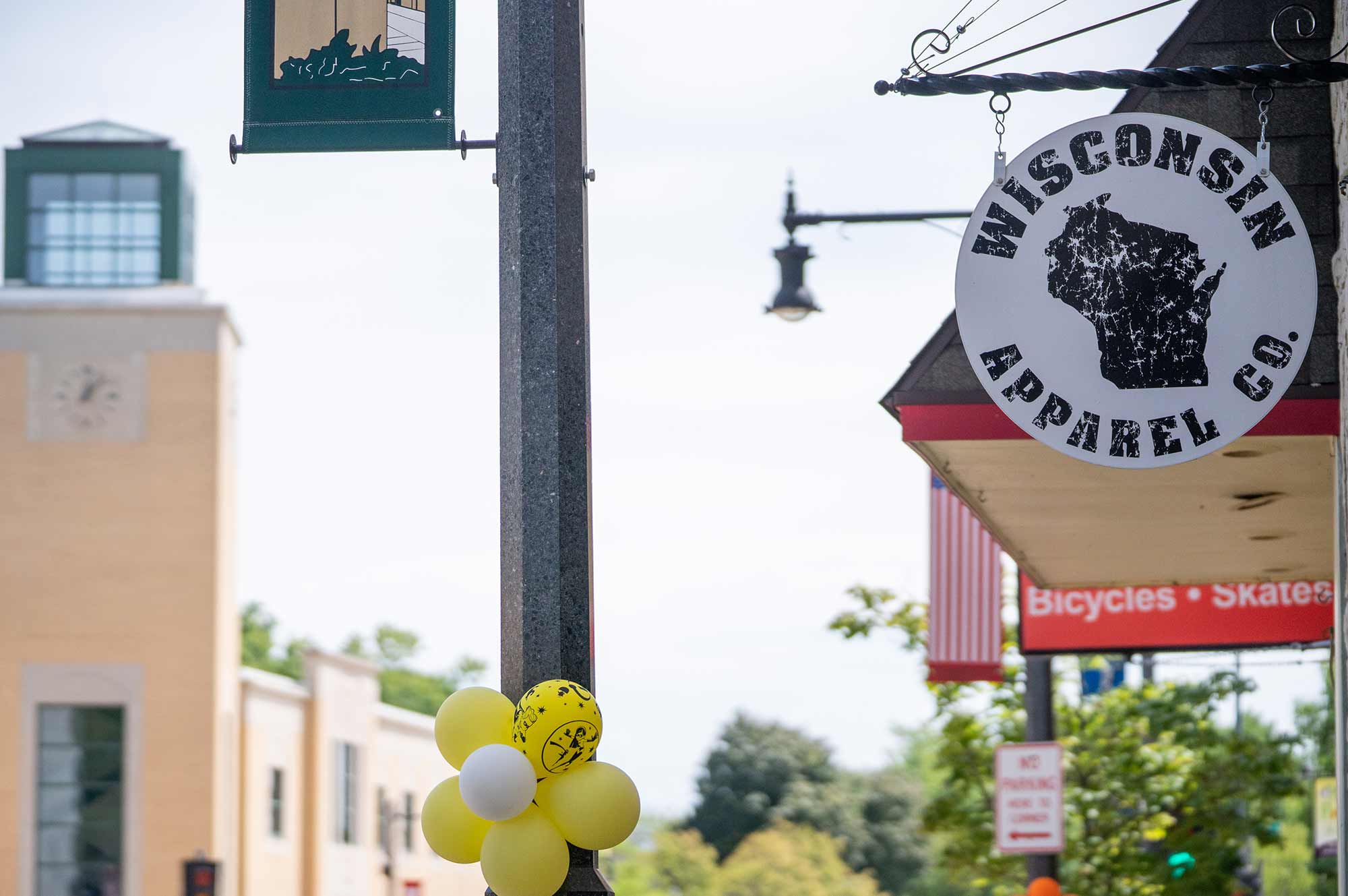 Street view with a Wisconsin Apparel Co. sign, yellow and white balloons on a pole, and a building with a clock tower in the background.
