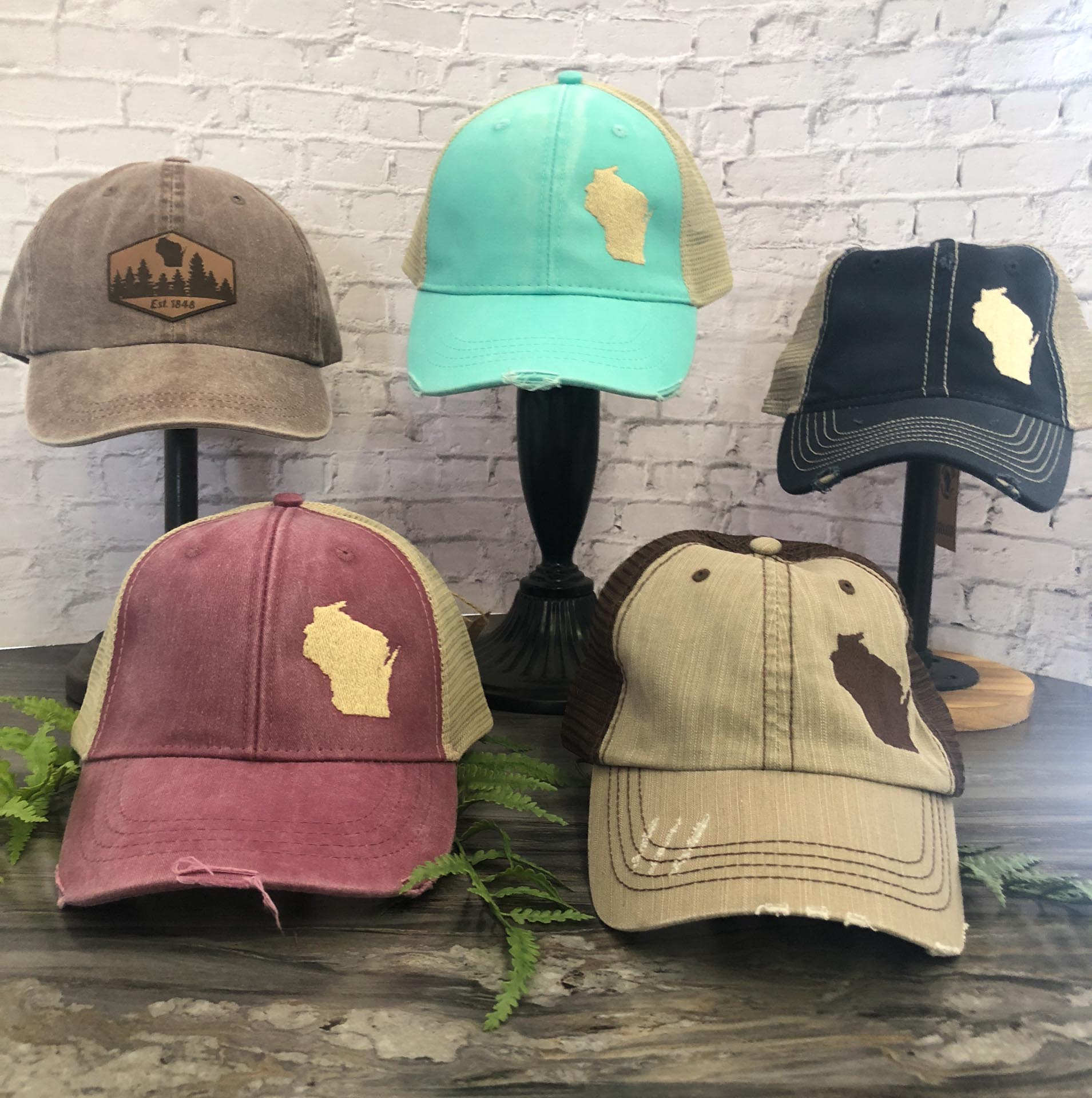 Five baseball caps with a Wisconsin state outline design are displayed on stands and a wooden surface against a white brick background.